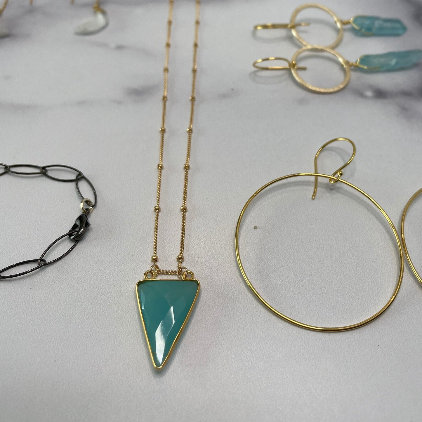 aqua chalcedony arrowhead pendant hanging on hold beaded necklace on gold hoop next to gold hoops and other gems on white marble table