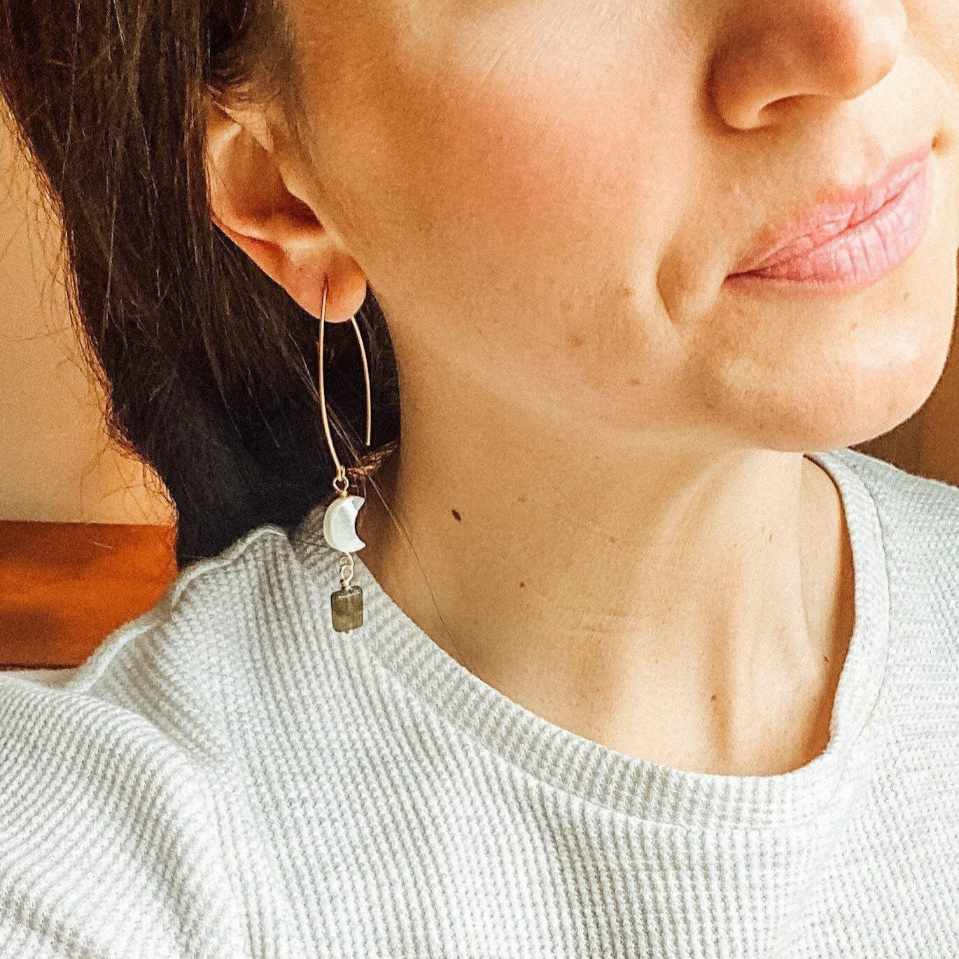 two gold earrings with mother of pearl gem and labrodorite gems hanging on gold metal hanging on girl with brunette hairs ear while wearing a white shirt