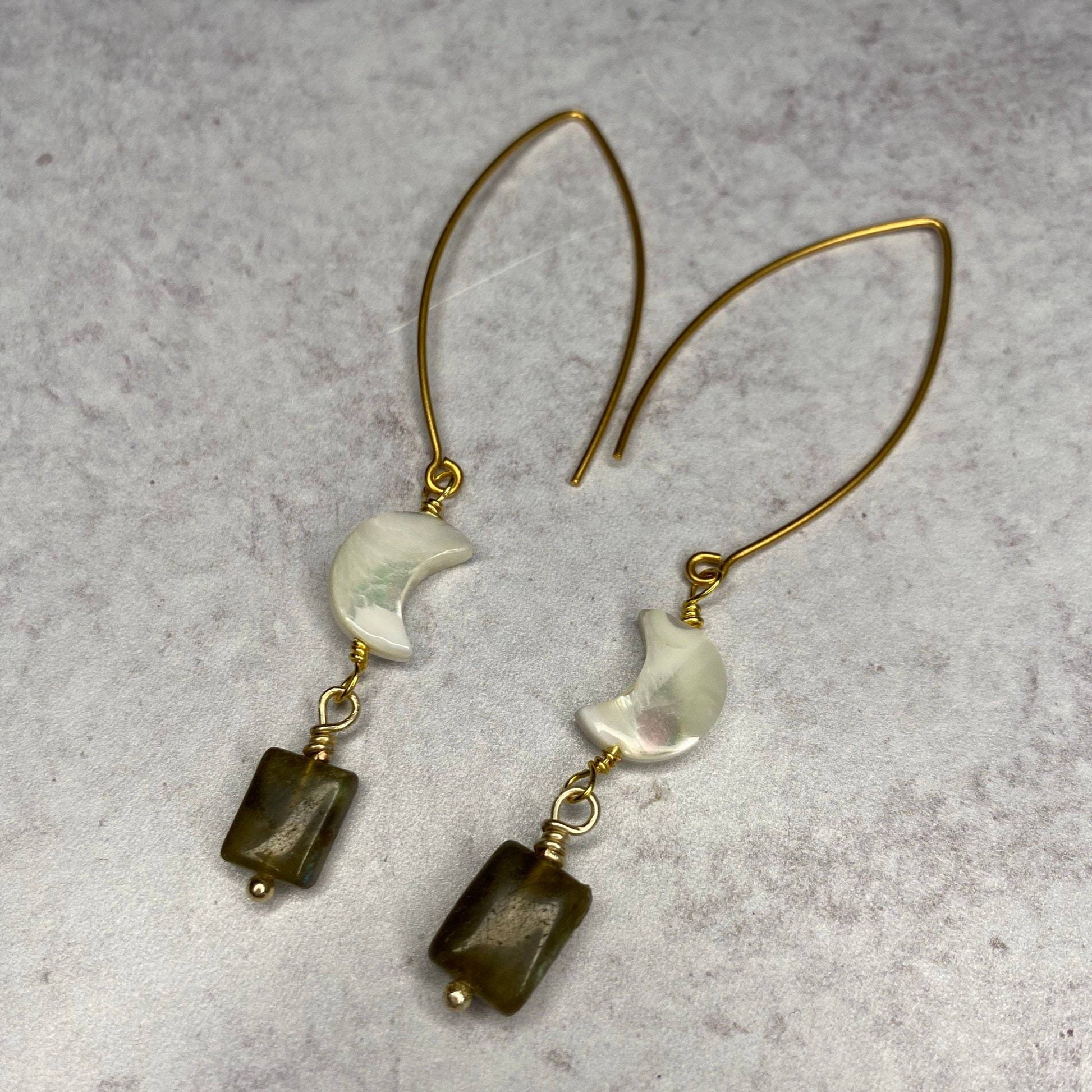 two gold earrings with mother of pearl gem and labrodorite gems hanging on gold metal with white stone background