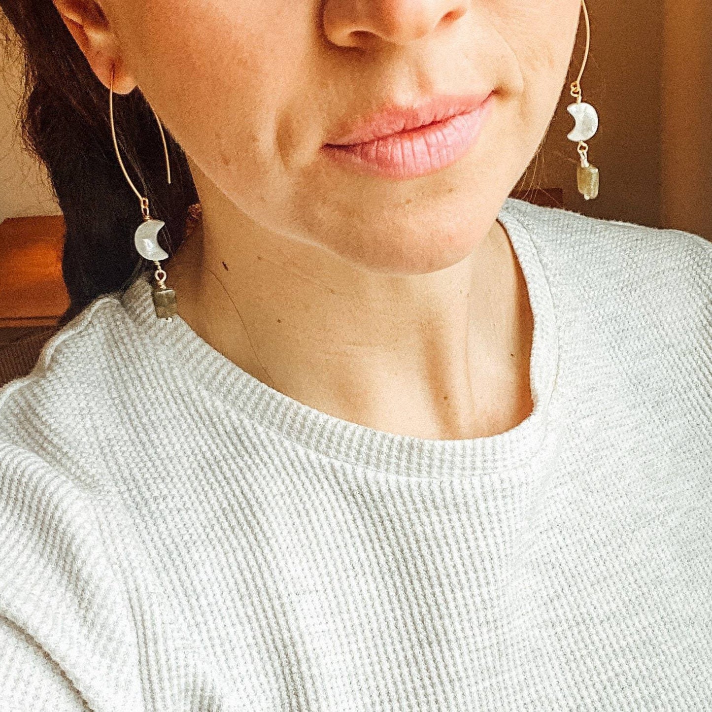 two gold earrings with mother of pearl gem and labrodorite gems hanging on gold metal hanging on brunettes ears while wearing a white shirt and pink lipstick