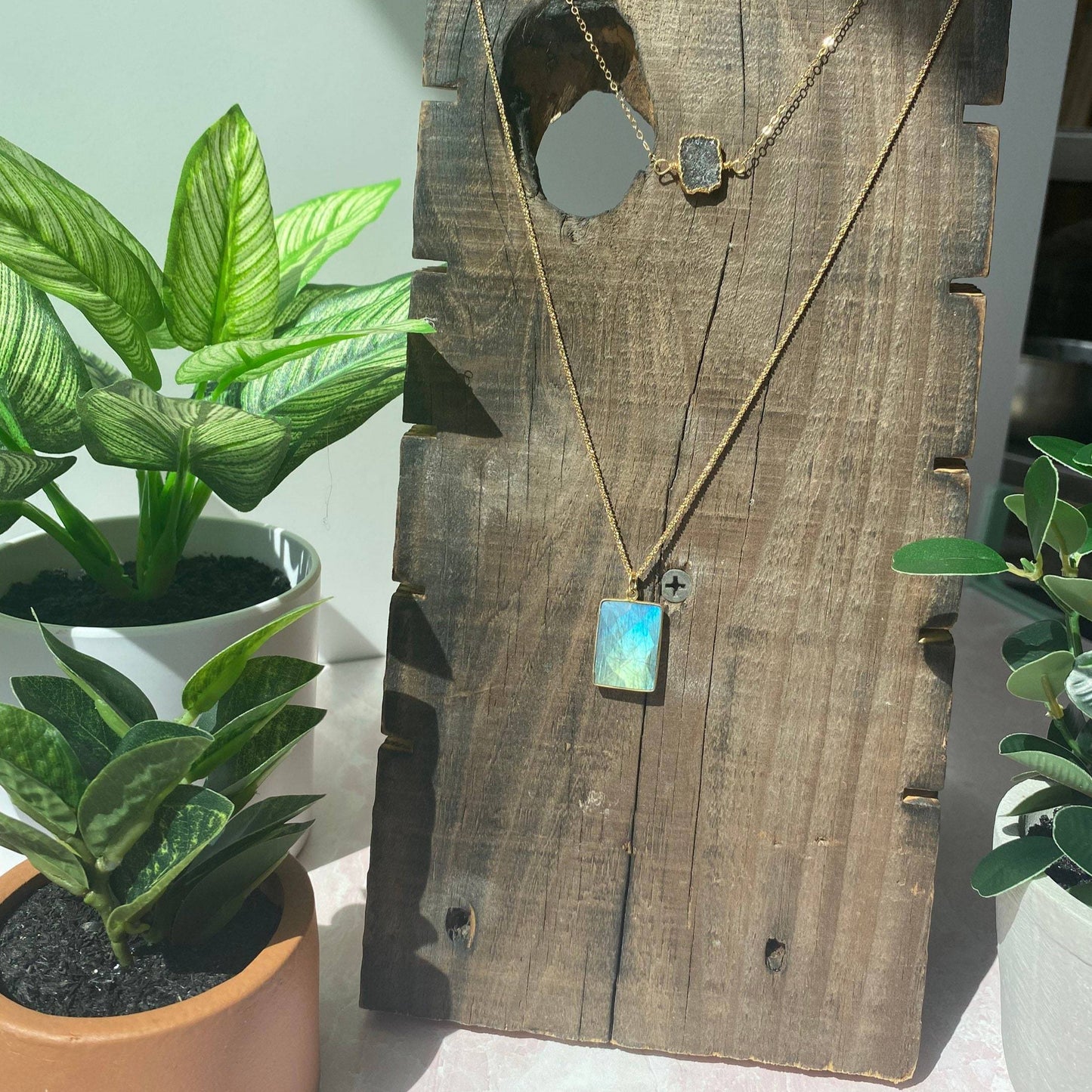 beautiful blue square labrodorite bezzel pendent hanging on gold chain hanging on reclaimed wood piece with three green plans in two white and one tan planter