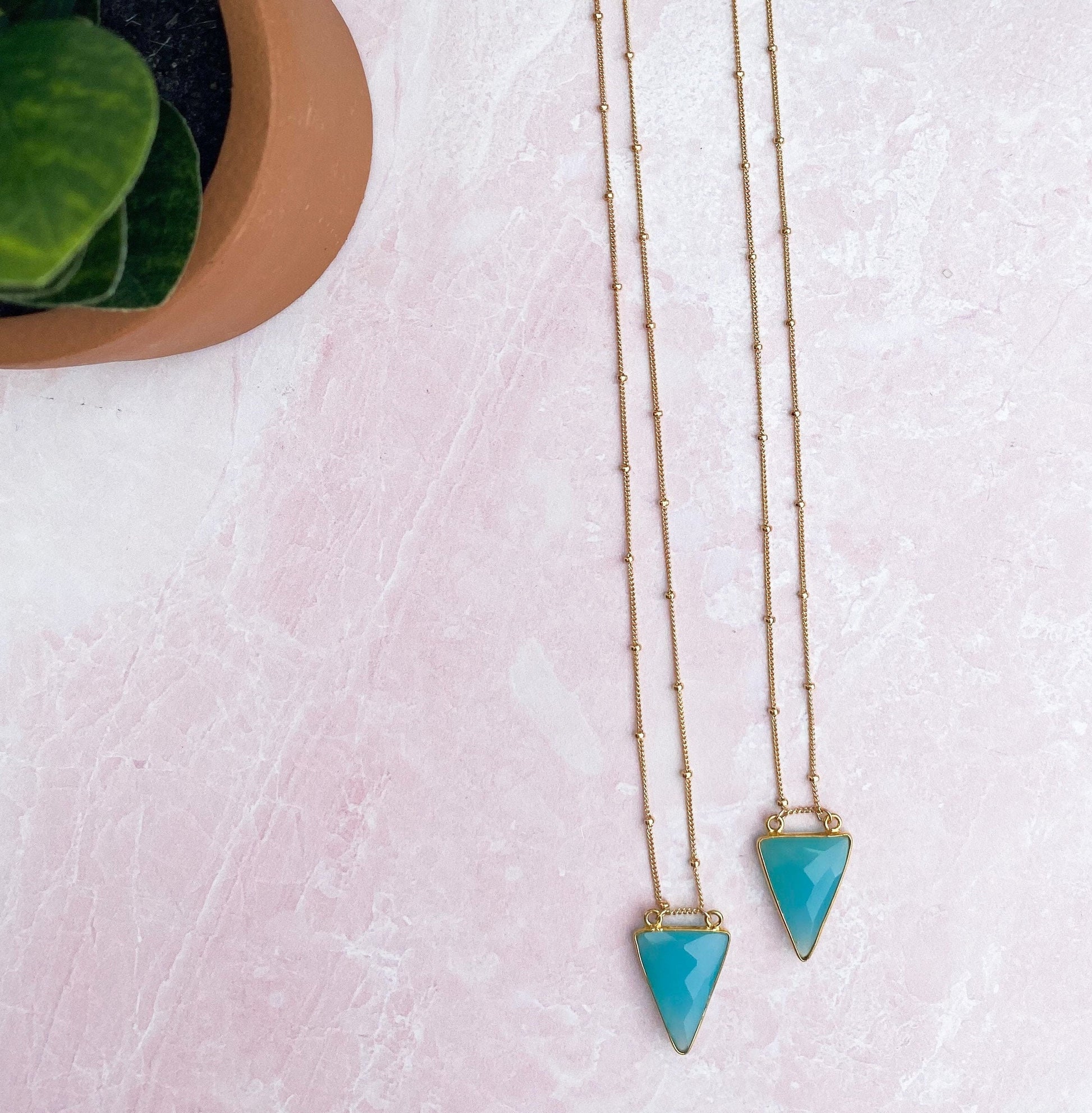 aqua chalcedony arrowhead pendant hanging on hold beaded necklace on pink quartz table along with green plant in tan bowl