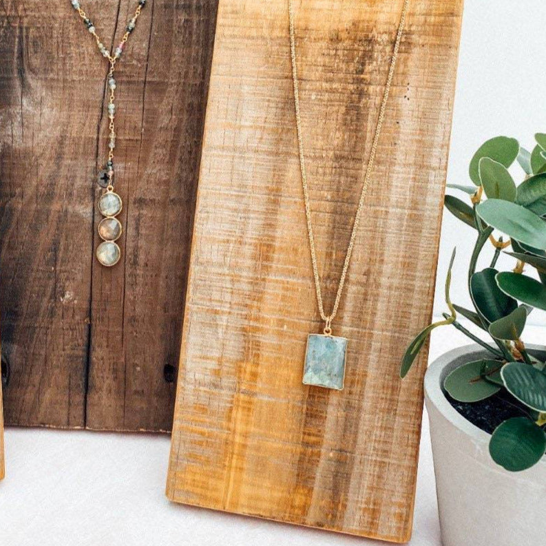 three gem neckace on reclaimed wood next to beautiful blue square labrodorite bezzel pendent hanging on gold chain and reclaimed wood