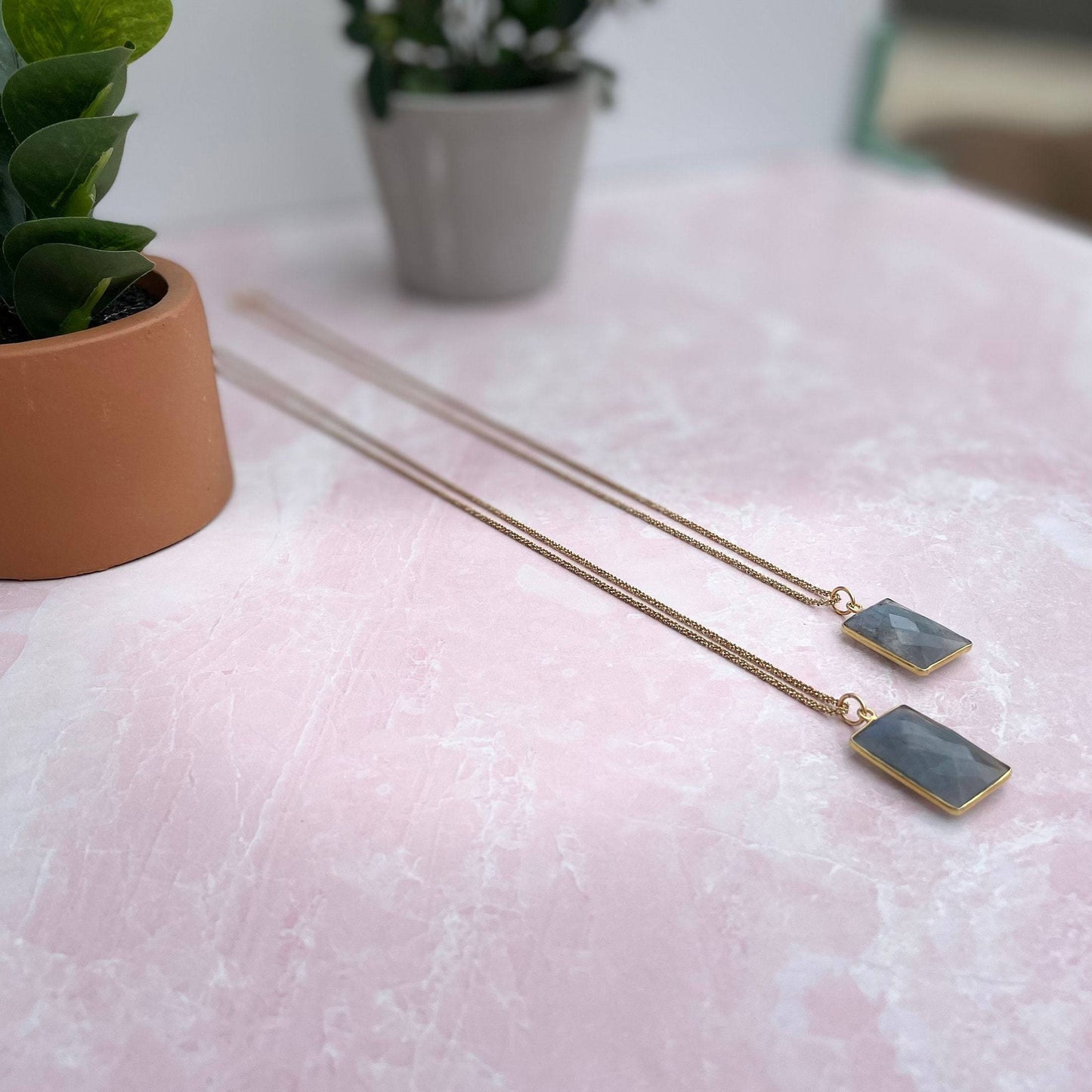 beautiful blue square labrodorite bezzel pendent hanging on gold chain on white quartz slab and two plants in tan and grey planter