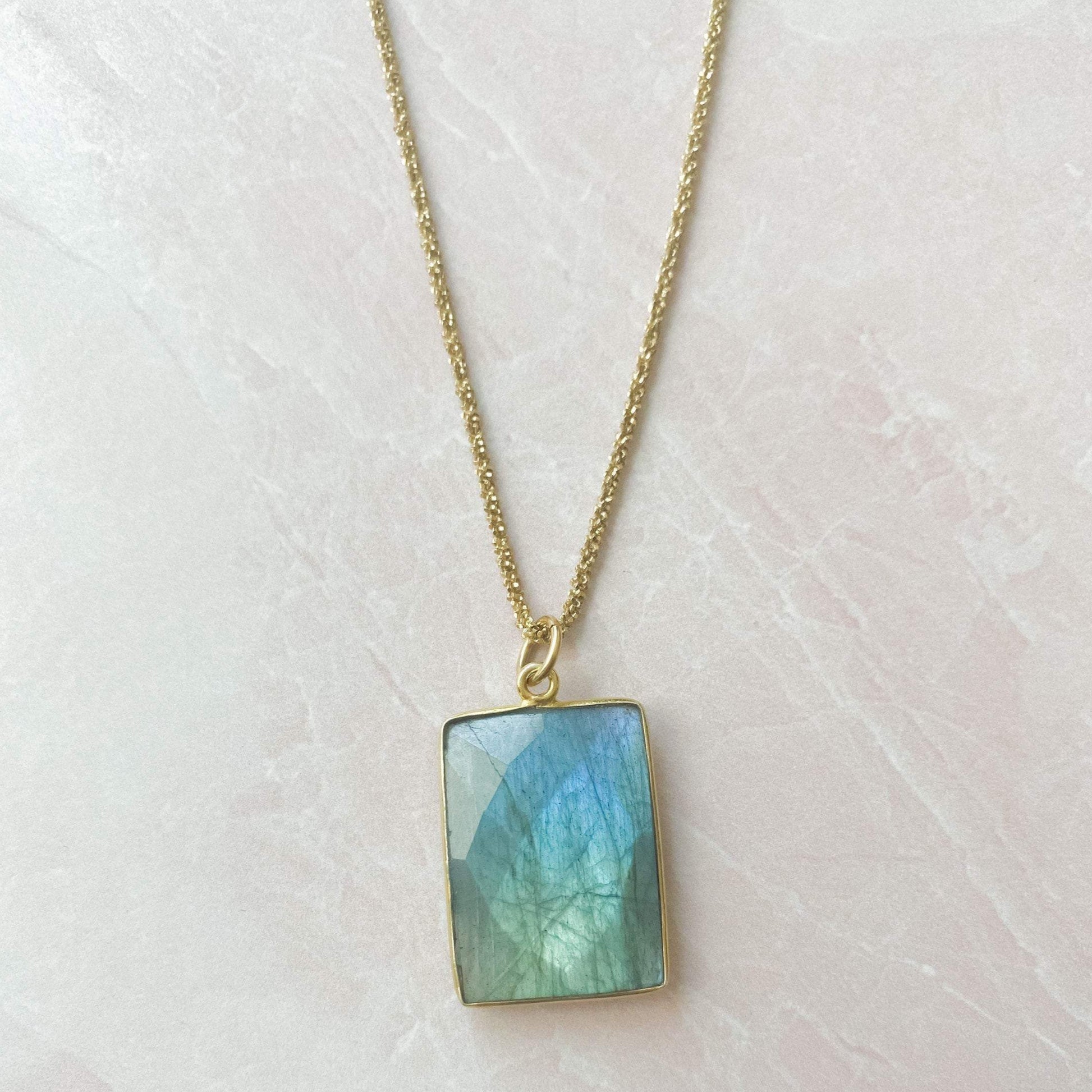 beautiful blue square labrodorite bezzel pendent hanging on gold chain and white marble slab