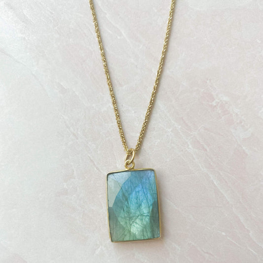 beautiful blue square labrodorite bezzel pendent hanging on gold chain and white marble slab